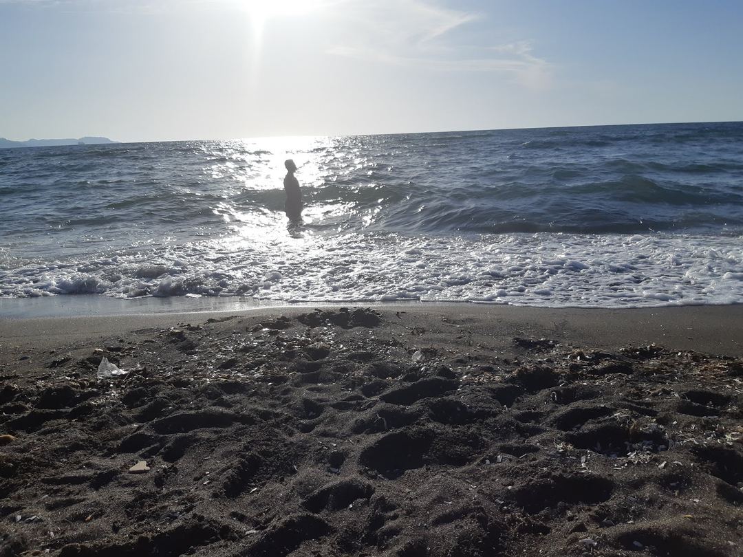 A person wades into the Mediterranean Sea at sunset with golden light reflecting off the water and rocky beach in the foreground.
