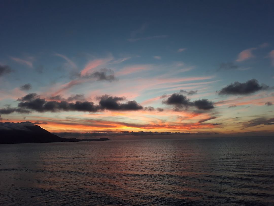 A dramatic sunset over the sea with mountains silhouetted against an orange and pink sky with scattered clouds.