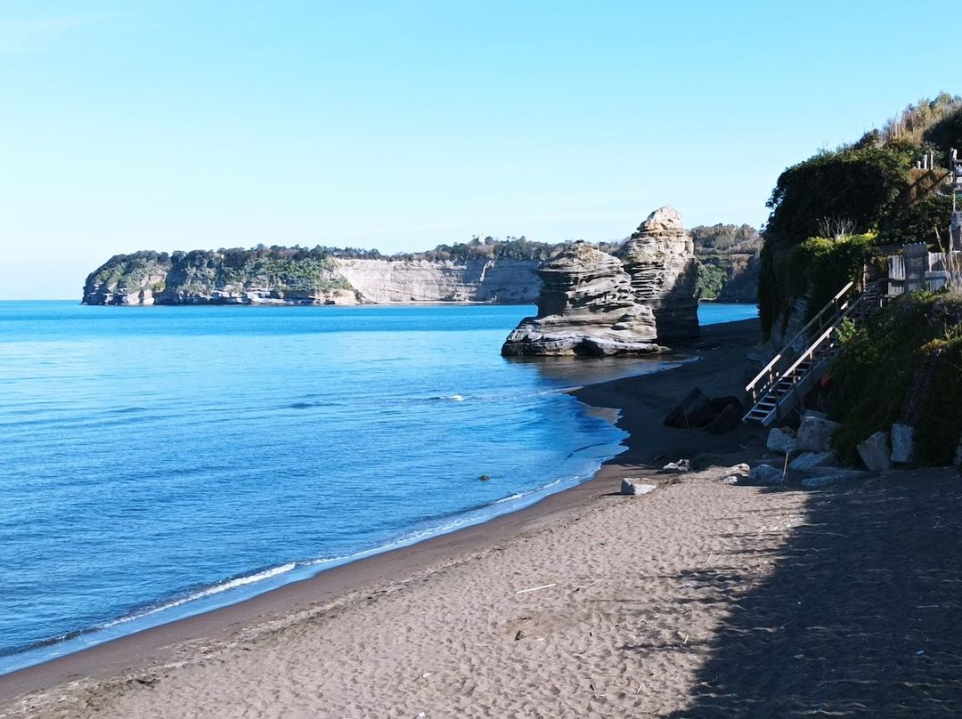A scenic beach cove with turquoise waters, dark sand, dramatic rocky cliffs, and lush vegetation overlooking the Mediterranean coast.